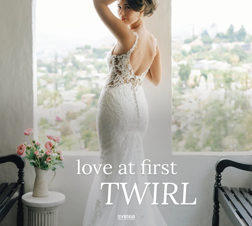 Stella York bridal gown SY8168, a mermaid wedding dress featuring an illusion lace bodice, deep open back, and intricate all-over lace and tulle skirt. The bride stands in a window overlooking a sunny cityscape.